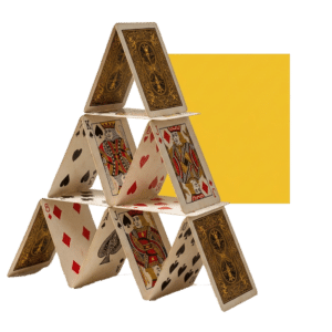 A three-tiered house of cards is built from an assortment of face and back-facing playing cards. The cards feature a classic, intricate gold, brown, and black design. In the second row, a red and gold Jack of Diamonds and a Jack of Hearts face outwards. In the bottom row, a Red Jack of Diamonds and an Ace of Spades face outwards. A large yellow square sits behind the cards to the right, against a plain white background. The image is a photograph taken from a slightly low angle, making the structure feel large. It is lit with soft, direct lighting.