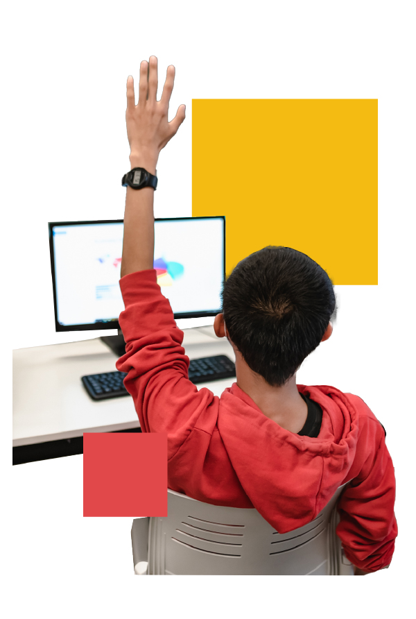 A student wearing a red hoodie sits in a classroom, raising their hand while looking at a computer screen displaying a colorful diagram. The student is seated on a white chair, with a keyboard and monitor on the desk in front of them.