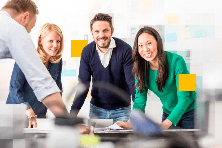 A medium-shot photograph of four smiling colleagues, three standing and one sitting at a table, in a modern-style open-plan office. The colleagues are standing around a collaborative table that has some blurred objects in the foreground, while in the background there is a whiteboard or corkboard wall with some blurred sticky notes or cards that are also partially obscured by two large solid yellow rectangle overlays.