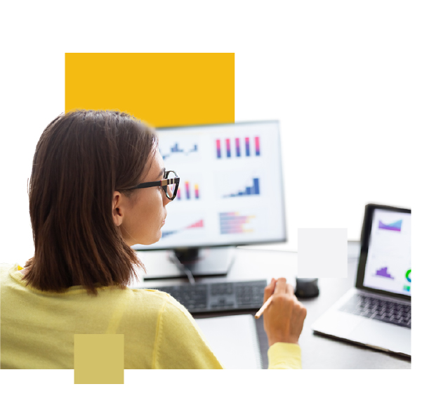 A woman is analyzing data on a computer screen with bar and line charts. She holds a pen while working at a desk with a laptop and an additional monitor displaying graphs.